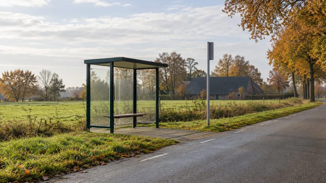 Leuge bushalte met waanden van glas en ne hoolten baanke langs ne weg boetn et doarp, met ne weide, hearfstbeume en 'n spil op de achtergroond.